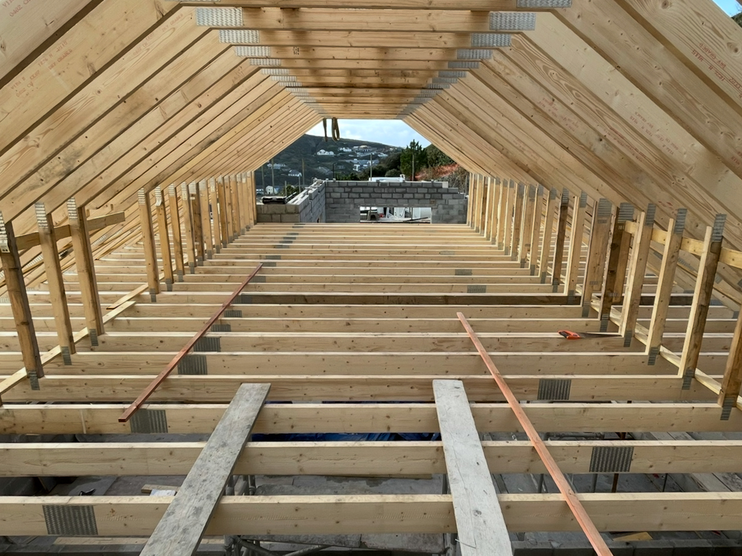 Interior view of a wooden pitched roof frame under construction with parallel rafters and floor joists, overlooking a hillside neighborhood